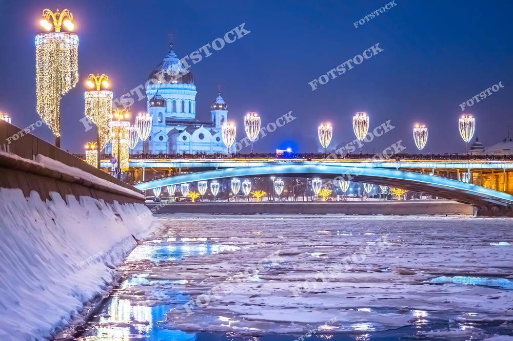 На фотографии изображён Храм Христа Спасителя, освещённый в зимнюю ночь. На переднем плане — покрытая льдом Москва-река и украшенный праздничной иллюминацией мост. Улицы и набережная оформлены световыми композициями и гирляндами. Атмосферу дополняют сверкающие фонари и снежные сугробы вдоль дороги. Вид подчёркивает торжественность и уют столицы в зимний период. Всё пространство наполнено светом и праздничным настроением.