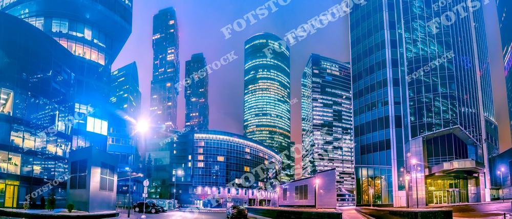 На фотографии изображён деловой район Москвы, который оживает в ночное время благодаря ярким огням и современным небоскрёбам. Высокие стеклянные здания отражают свет, создавая эффект сверкающего города. Панорама района показывает гармоничное сочетание современной архитектуры и городской инфраструктуры, подчёркнутой мягким светом, создающим атмосферу динамичного и современного мегаполиса.