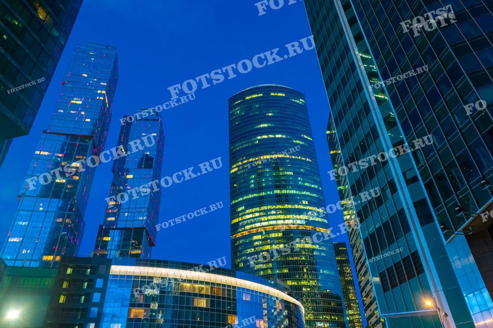 На фотографии изображены небоскребы делового центра Москва-Сити в ночное время. Освещение зданий подчеркивает современную архитектуру и высокие технологии, создавая впечатляющий городской пейзаж.