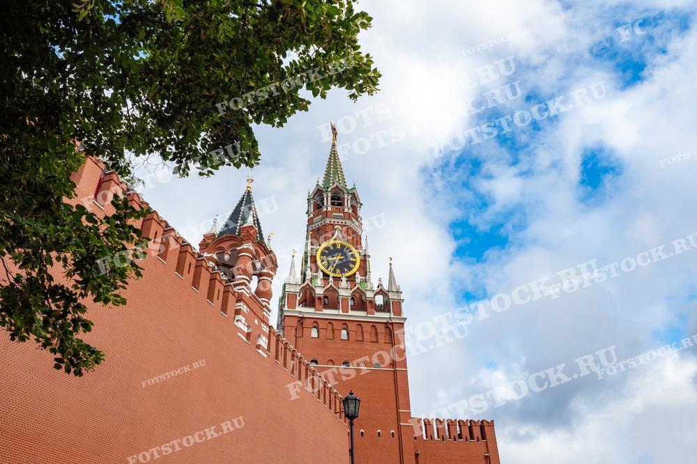 Фотография показывает Спасскую башню Московского Кремля с ее знаменитыми часами. На переднем плане видна красная кирпичная стена Кремля. Летний день с голубым небом и облаками добавляет яркости изображению. Ветви дерева обрамляют вид сверху слева, создавая приятный контраст с архитектурой.