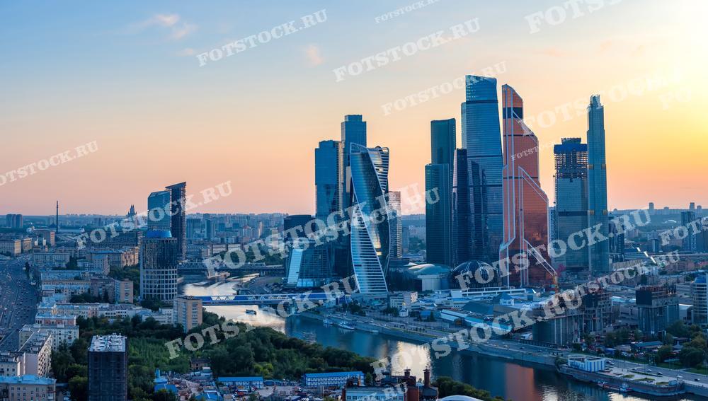 На фотографии изображена вечерняя панорама делового центра Москва-Сити и Москва-река. Закатное солнце отражается в стеклянных фасадах небоскребов, создавая живописный вид на современную Москву.