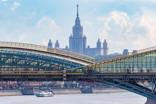 На фотографии изображен стеклянный пешеходный мост, простирающийся над рекой Москвой, а также высотное здание на набережной. Это фото иллюстрирует архитектуру столицы России и красоту пейзажа города. Вид на Москву представляет собой прекрасную панораму, которая может быть использована для туристических материалов о Москве, городских экскурсий и рекламы столичных достопримечательностей.