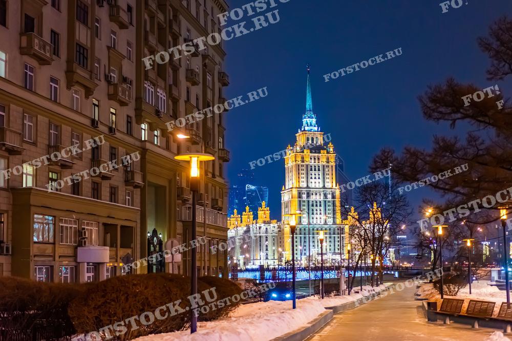 На фотографии изображена одна из высоток Москвы — отель Украина. Здание освещено, его золотистые огни контрастируют с ночным голубым небом, создавая живописную и теплую атмосферу. Дорожные фонари и следы машин на снегу добавляют уют и динамичность сцене. Это изображение подойдет для использования в публикациях о московской архитектуре, туристических проспектах или как часть декоративного оформления в статьях о путешествиях по России.
