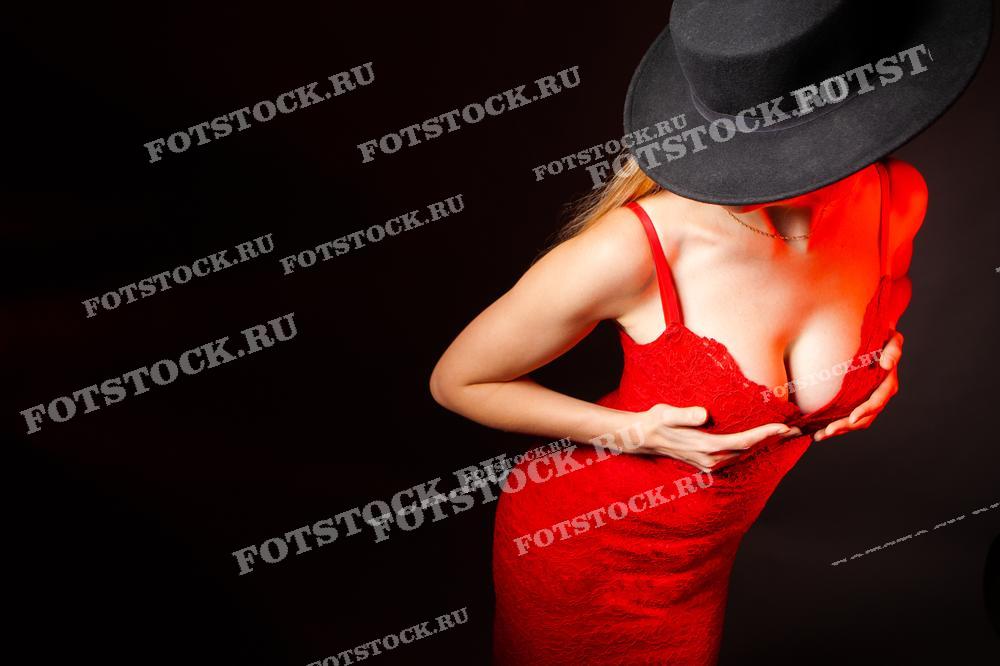 На изображении женщина в красном кружевном платье и чёрной шляпе, находящаяся в откровенной позе на фоне тёмного фона. Фотография подчеркивает загадочность и чувственность, создавая эффект драматического освещения и акцента на силуэте.