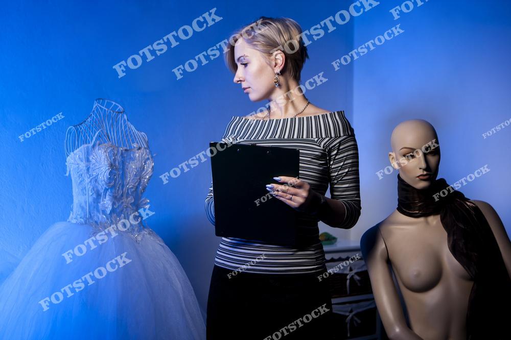 Фотоснимок показывает девушку — fashion-дизайнера, которая делает заметки и работает над пошивом свадебных платьев, где изображение передаёт творческий и профессиональный процесс в ателье. Эта фотография выглядит как рабочая иллюстрация модельера, а картинка создаёт аккуратный и вдохновляющий слайд создания свадебной моды.