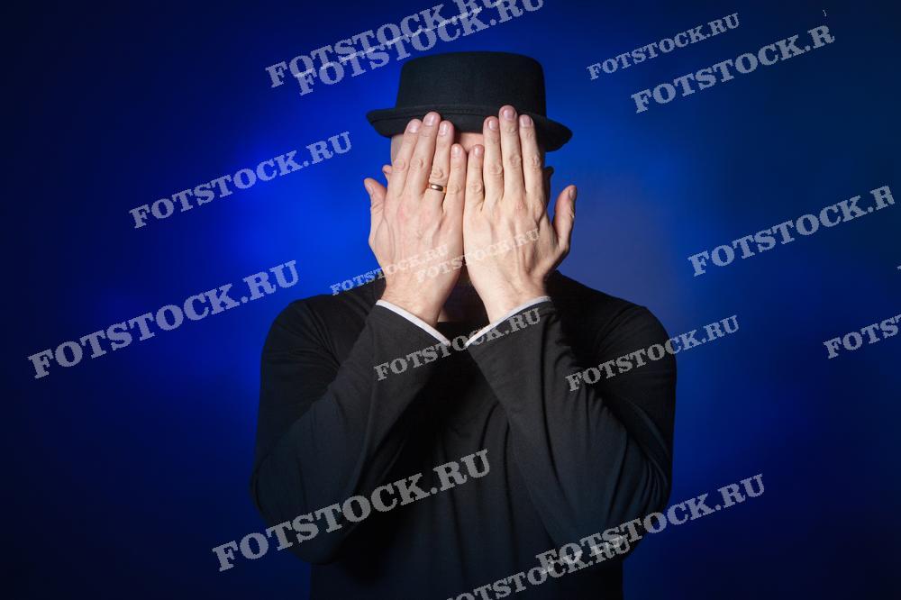 На изображении мужчина в чёрной шляпе закрывает лицо руками на фоне глубокого синего цвета. Фотография представляет собой концептуальный портрет, передающий ощущение таинственности и интриги.