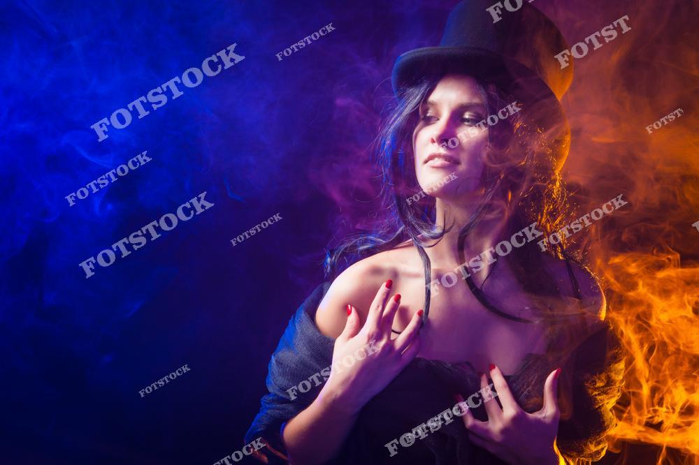 Фотография женщины в черном наряде и цилиндре, окруженной клубами фиолетового и оранжевого дыма. Контрастные цвета и освещение создают атмосферу загадочности и магии. Образ сочетает элегантность, чувственность и мистический стиль.