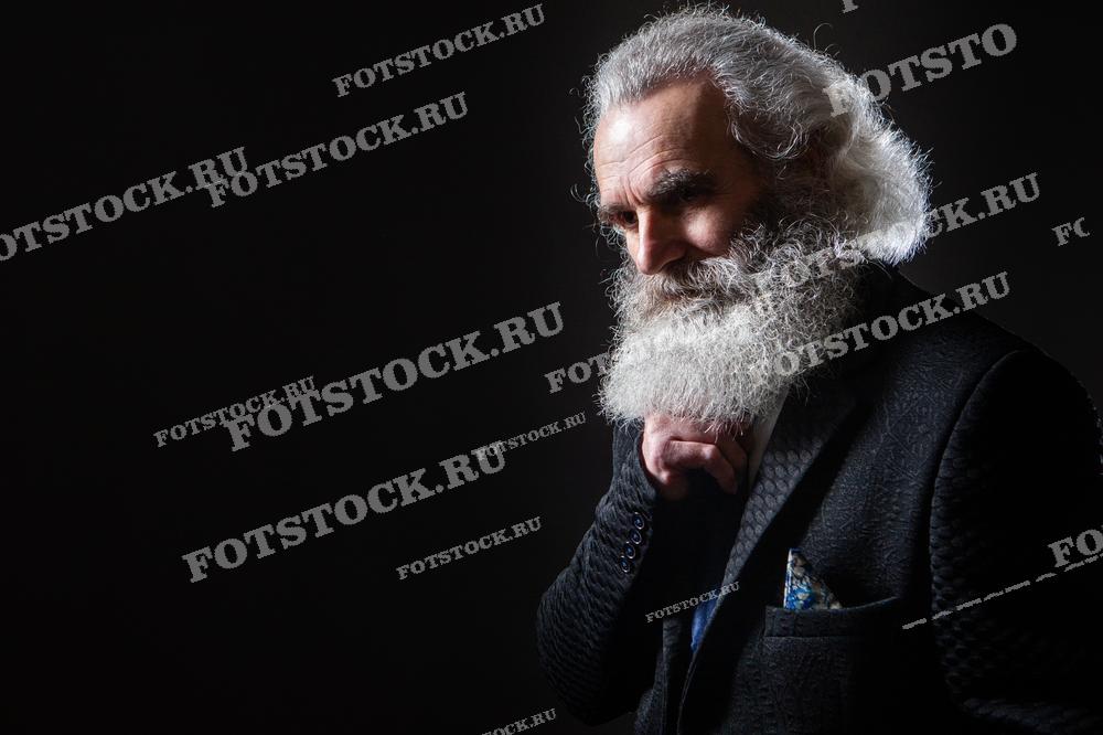 На изображении пожилой мужчина с густой седой бородой, одетый в тёмный костюм, позирует на фоне с приглушённым светом. Фотография передаёт ощущение мудрости, силы характера и достоинства, подчёркивая зрелость и опыт.