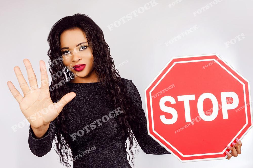 На фотографии изображена молодая женщина с длинными вьющимися волосами, которая держит в руках красный знак "STOP" и одновременно показывает ладонь вперёд, как сигнал "остановись". Это студийный снимок на белом фоне, символизирующий уверенность, защиту личных границ и социальные месседжи.