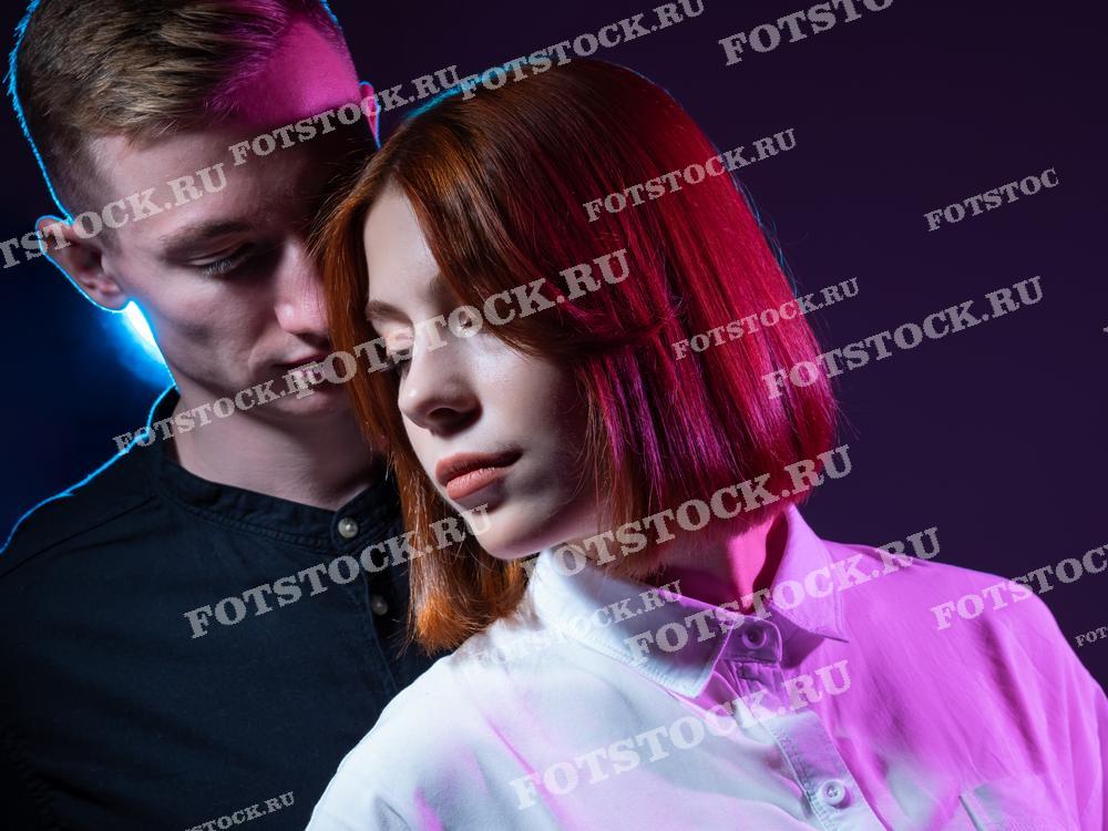 На фотографии изображена молодая пара. Разноцветное освещение создает атмосферу загадочности и романтики, подчеркивая эмоциональную связь между юношей и девушкой.