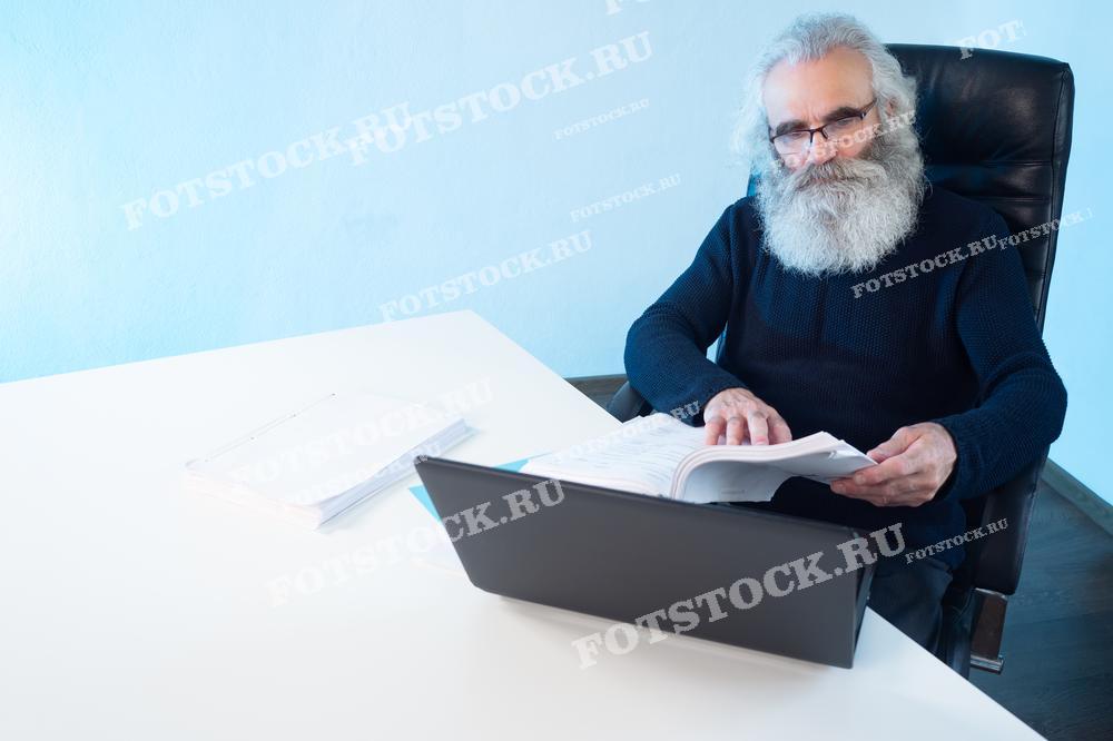 На фотографии изображен пожилой мужчина с густой седой бородой, сидящий за офисным столом и работающий за ноутбуком. Он просматривает документы, демонстрируя сосредоточенность в своей работе.
