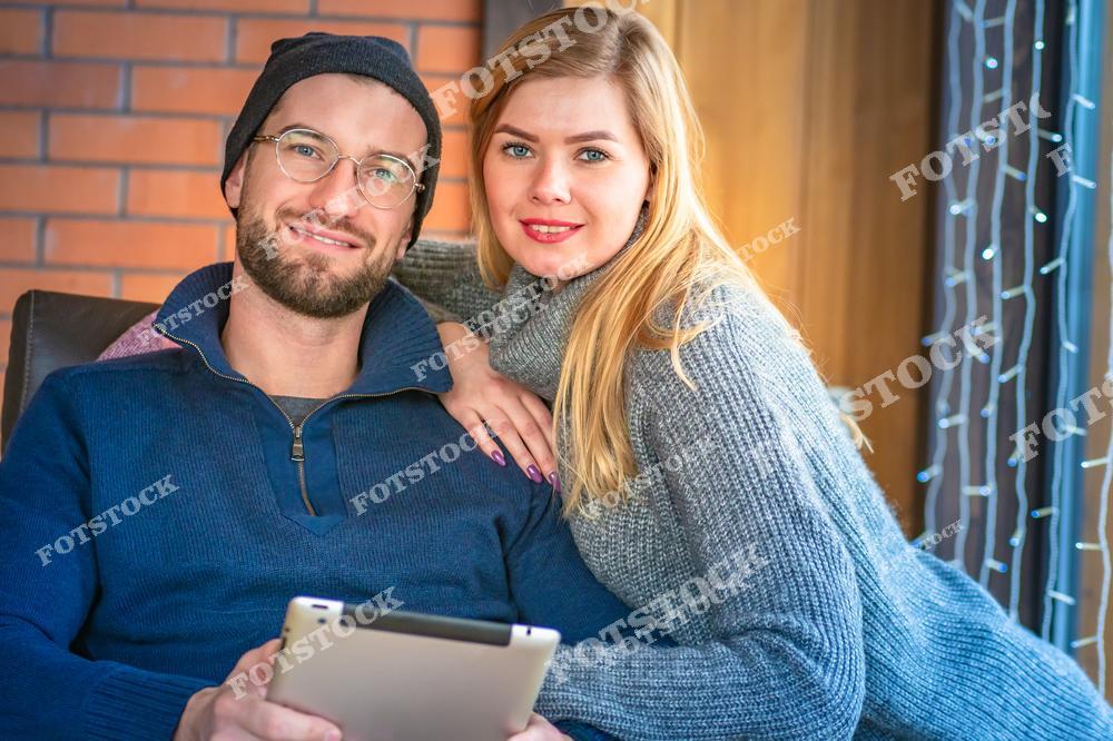 На фотографии изображена молодая пара в коттедже, сидящая на фоне кирпичной стены и деревянного интерьера. Девушка нежно опирается на плечо мужчины, который держит планшет. Их теплые улыбки создают атмосферу уюта и доброжелательности.