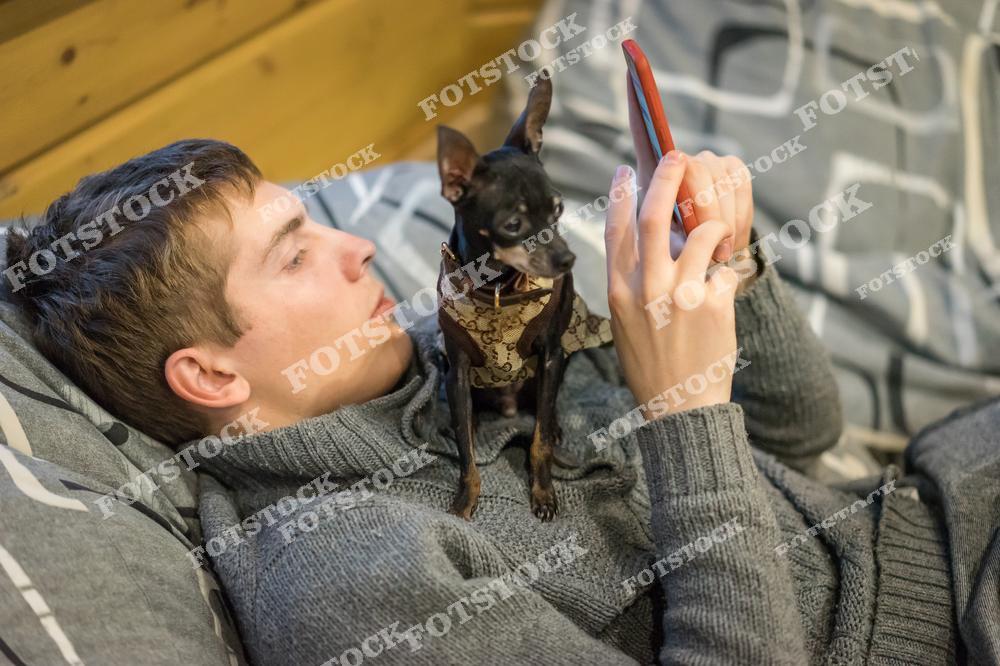 На фотографии молодой человек лежит на диване, увлеченно смотря в смартфон, а его маленькая собака сидит на груди. Окружающая обстановка создаёт уютное ощущение домашнего комфорта. Взаимодействие человека и животного символизирует теплое и доверительное отношение.