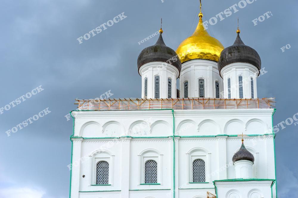На фотографии изображены золотые купола Троицкого собора в Пскове, которые выделяются на фоне темного неба. В данный момент идут реставрационные работы, подчеркивающие историческую ценность этого архитектурного памятника.