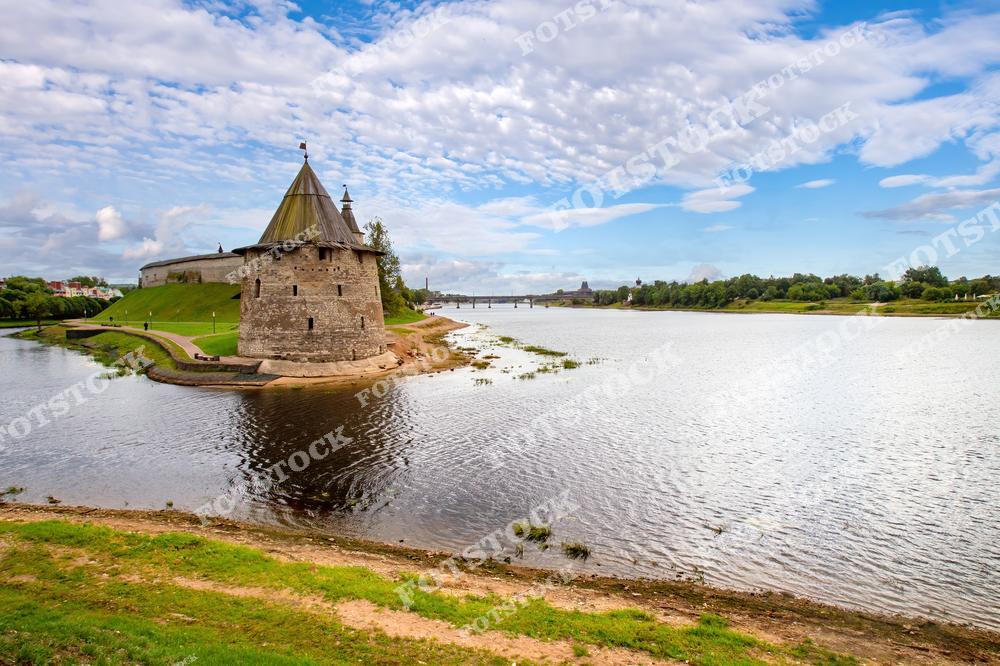 На фотографии изображена башня Псковского Кремля, расположенная на берегу реки Великой. Архитектурное наследие дополнено живописным пейзажем с голубым небом и спокойной водой. Вид передаёт гармонию истории и природы.