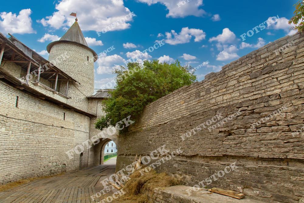 На фотографии изображена крепостная башня Псковского Кремля, окружённая массивными каменными стенами. Уникальная архитектура под голубым небом с облаками подчёркивает историческое значение объекта. Каменные сооружения создают атмосферу средневековья.