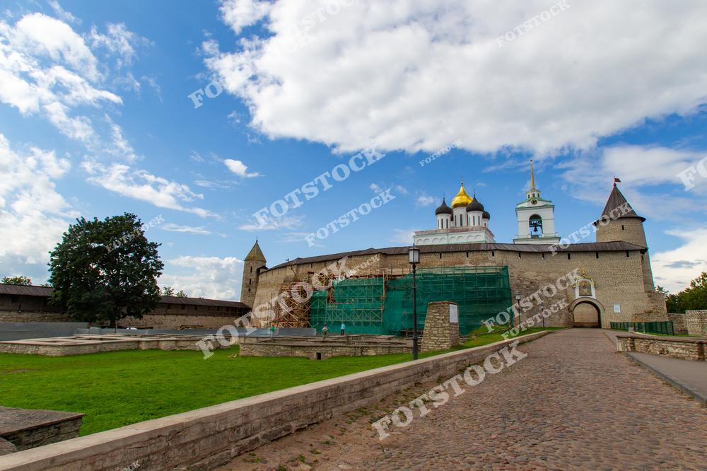 На фотографии изображен Псковский Кремль с Троицким собором, расположенный на возвышенности. Каменные стены и башни крепости гармонично сочетаются с зелеными лужайками и голубым небом. Этот памятник истории и архитектуры символизирует богатое наследие России.