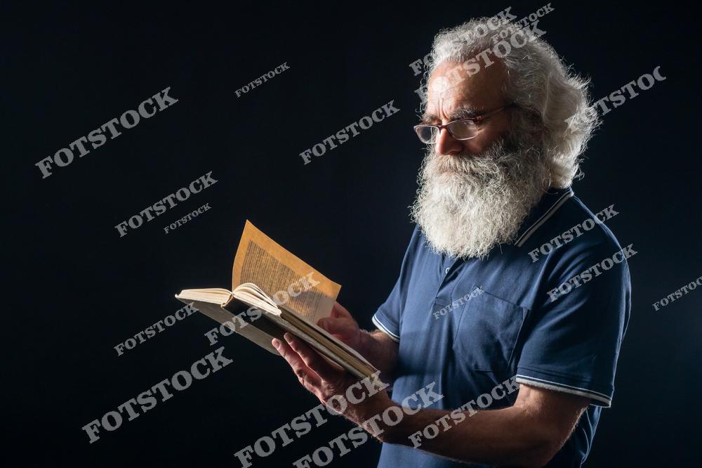 Пожилой мужчина с густой седой бородой и в очках держит открытую книгу. На фотографии изображен процесс чтения в спокойной и сосредоточенной обстановке. Темный фон подчеркивает сосредоточенность и внимание к чтению.