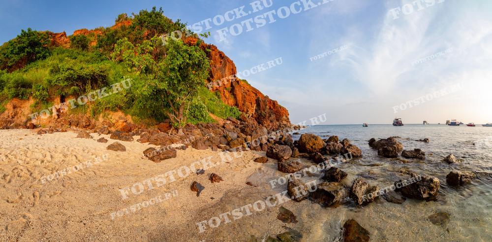 Панорамная фотография живописного пляжа с песком, камнями и скалистыми холмами, покрытыми зеленью. Море спокойное, на горизонте видны лодки. Этот тропический пейзаж Таиланда сочетает в себе природную красоту пляжа и спокойствие моря, создавая атмосферу отдыха и умиротворения.
