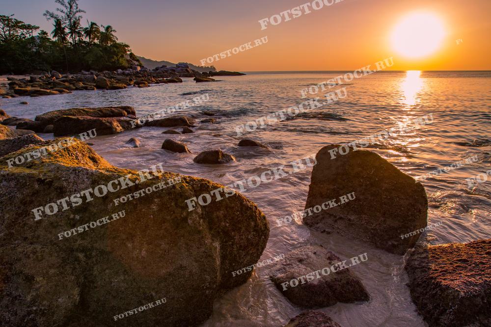 Фотография закатного солнца над морем на фоне каменистого пляжа в Таиланде. Тёплые лучи заката освещают воду и скалы, создавая атмосферу спокойствия и умиротворения. Это изображение подчеркивает природную красоту побережья и гармонию природы.