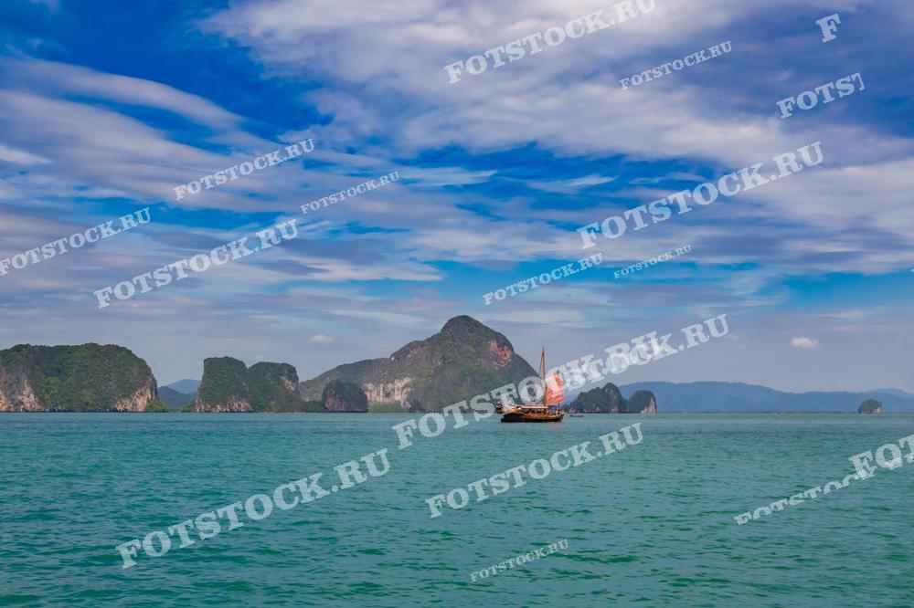 На фотографии традиционная парусная лодка плывёт по спокойным водам залива Пханг Нга, Таиланд. Лодка окружена тропическими островами, а облачное небо добавляет глубину пейзажу. Эта сцена передаёт ощущение уединённости и гармонии с природой.